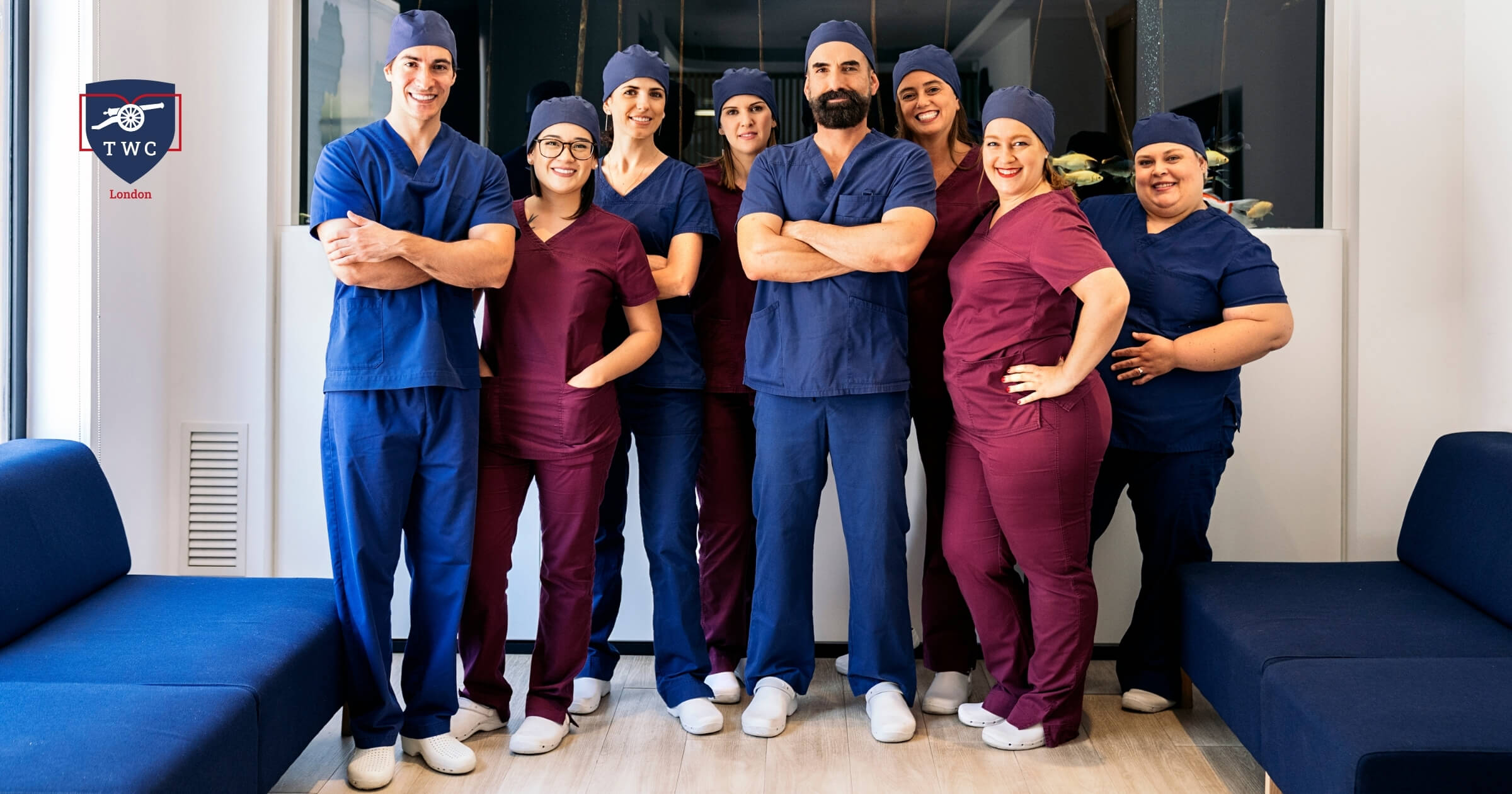 A group of doctors and nurses stands together in front of a blue couch, smiling and dressed in medical attire.